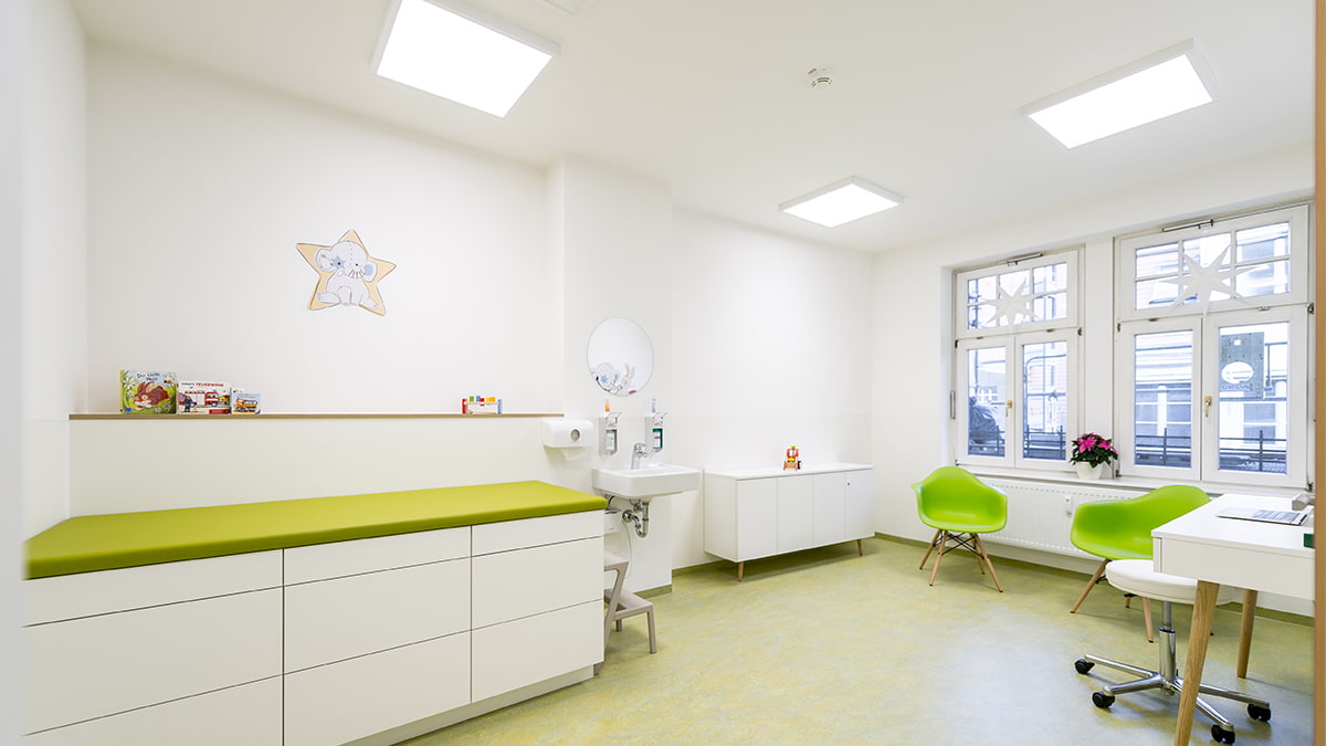 Kinderarztpraxis Hussack Leipzig Blick in großen Behandlungsraum mit Wandbildern, Ablagefläche, grünen Akzenten und zwei großen Fenstern