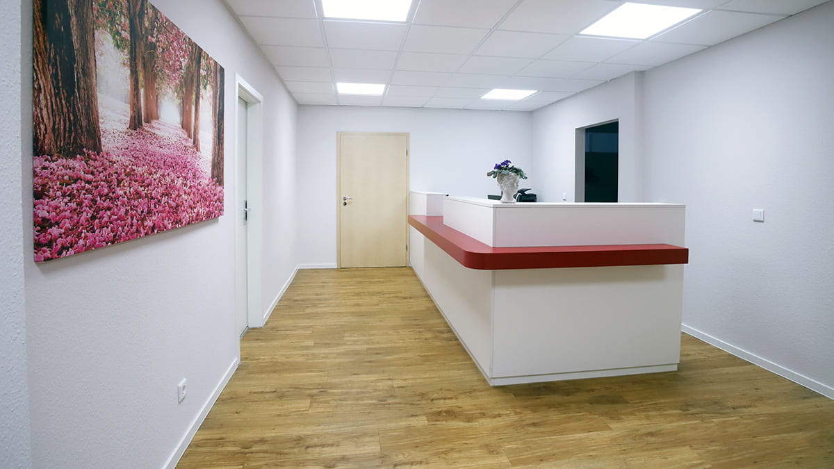 Referenz Praxis für Allgemeinmedizin Altenburger Blick auf den Empfangsbereich in moderner, weißer Optikmit einer Ablage in Rot, einem Boden in Holzoptik sowie einem Wandbild mit rosafarbenen Blumen in einer Allee