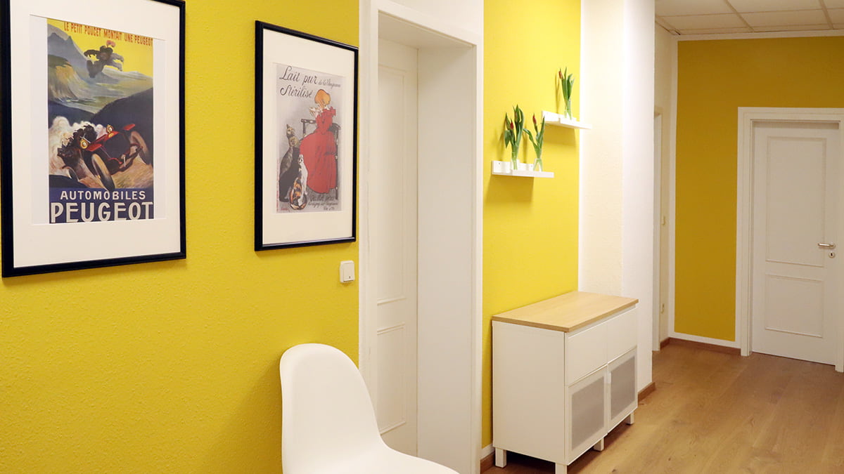Referenz Praxis für Psychotherapie Peterhänsel Leipzig Blick von der Eingangstür in Richtung der Sprechzimmer mit gelber Wandfarbe, Möbeln in weißer Farbe und Bildern sowie Blumen an der Wand