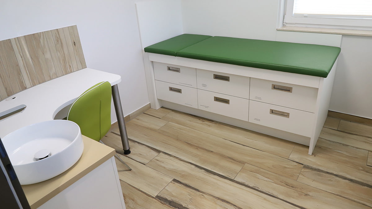 Antonius-Apotheke Ilmenau Behandlungszimmer mit einer Liege mit grüner Auflage & engebauten Schubladen darunter sowie einem abgerundeten Tisch mit einem Waschbecken & einem grünen Stuhl davor